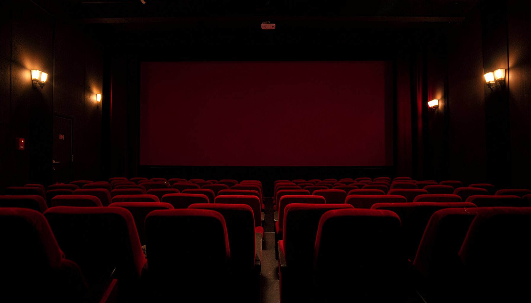 Darkened cinema auditorium with rows of red velvet seats, warm ambient lighting, and a large blank screen at the front, evoking a sense of anticipation and storytelling tradition.